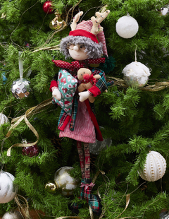 Muñeca de Mrs. Claus vestida con pijama de rayas y bata, sosteniendo una almohada y un peluche de reno, decorando un árbol de Navidad.