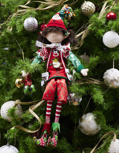 Duende navideño con bermudas y balón de fútbol, decorado con elementos festivos, colgado entre ramas de un árbol de Navidad.