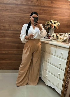 Mulher posando em um espelho, vestindo uma blusa branca e calças largas de cor bege, com um fundo de parede de madeira e uma mesa decorada. Pantalona nude, calça pantalona, pantalona, calça larga, calça cor clara, calça nude, 