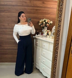 Mulher posando em frente a um espelho, vestindo uma blusa branca com detalhes e calças largas azuis, com um fundo de madeira e um vaso de flores ao lado. Calça pantalona na cor azul marinho, calça azul marinho, pantalona