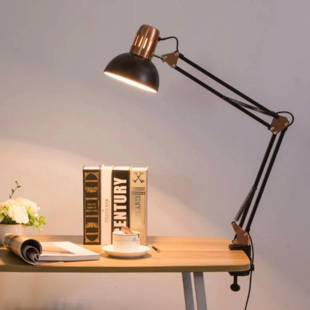 Uma luminária de mesa com braço articulado, iluminando um ambiente de trabalho com livros e uma xícara de café.