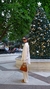 Mujer vestida con un conjunto de seda blanco, de pie junto a un árbol de Navidad decorado, sosteniendo un bolso marrón.