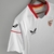 Camisa branca do Sevilla FC 22/23, modelo torcedor Castore, com detalhes em vermelho e patrocinadores visíveis.

