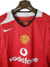 Camisa retrô do Manchester United de 2005, na cor vermelha com detalhes em preto e branco, com logo da Nike e patrocínio da Vodafone.

