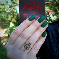 Imagem de uma mão com unhas verdes, usando um anel de aço inoxidável com design de nó de bruxa, segurando um cartão com símbolos místicos ao fundo.