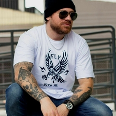 Homem com tatuagens usando a camiseta branca modelo Fly With Me da marca LK-LIKE, sentado em um corrimão.