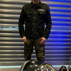 Homem posando em frente a uma parede metálica, vestindo uma jaqueta de sarja premium, edição limitada da marca LK-LIKE, e calças camufladas, com uma motocicleta ao fundo.