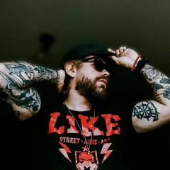 Homem com tatuagens, usando óculos escuros e uma camiseta preta clássica da marca LK-LIKE. Ele está posando com a mão na cabeça.