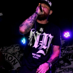 Homem com barba e tatuagens cantando em um palco, usando uma camiseta preta da marca LK-LIKE com a inscrição "NEW YORK".