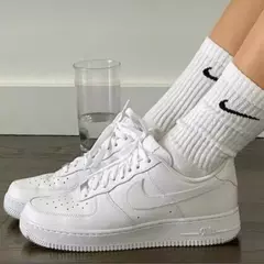 Um par de tênis Nk Air Force 1 brancos, usados com meias brancas Nike, em um piso de madeira, ao lado de um copo de água.