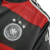 Detalhe da camisa retrô da Seleção da Alemanha II 2014, com cores preta e vermelha, exibindo o emblema da Deutscher Fussball-Bund e três estrelas.