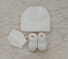 Conjunto de três peças para bebês com gorro, luvas e sapatinhos brancos sobre um fundo claro.