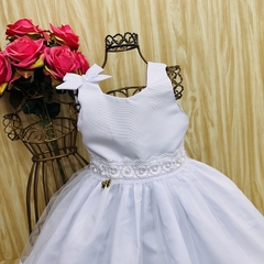 Vestido de festa infantil Elida, branco, com detalhes em pérolas e laço na alça, sobre um fundo claro.