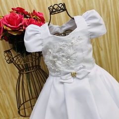 Vestido de festa infantil Analuz em tom branco, com detalhes em renda e laço na cintura, exibido em um cabideiro decorativo ao lado de flores rosas.