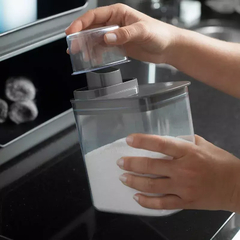 Pessoa utilizando um dispenser com medidor para dosar pó em um recipiente transparente.