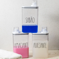 Três dispensadores transparentes com rótulos 'Sabão', 'Amaciante' e 'Alvejante', dispostos um sobre o outro, com líquidos coloridos dentro.