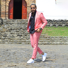 Hombre con traje premium color rosado, camisa negra y zapatillas blancas, posando en una calle empedrada.