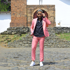 Hombre posando con un traje premium color rosado y camiseta negra, frente a una pared de ladrillo y escalinatas de piedra.