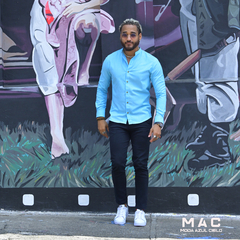 Hombre de pie frente a un mural colorido, vistiendo una camisa de lino azul claro y pantalones oscuros, con zapatillas blancas.