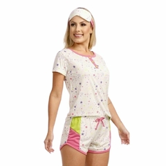 O pijama curto feminino na imagem tem uma blusa branca com estampas de estrelas coloridas e um short rosa. A blusa tem um detalhe de acabamento em rosa ao longo do decote e um laço rosa na frente. O short do pijama é predominantemente rosa, com uma faixa 