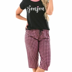 A parte superior do pijama é uma camiseta preta com mangas curtas. Na camiseta, a palavra “Bonjour” (bom dia, em francês) está escrita em letras cursivas brancas, acompanhada por um pequeno coração rosa. A parte inferior do pijama consiste em calças xadre