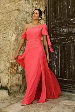 Modelo vestindo um vestido de festa rosa, com um ombro à mostra, em um cenário rústico.