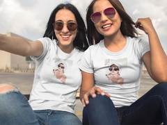 Duas mulheres sorrindo usando camisetas brancas com estampa de rostos, posando para a câmera.