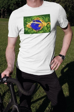 Homem usando uma camisa branca com estampa colorida inspirada na bandeira do Brasil, posando ao lado de uma bicicleta em um gramado.