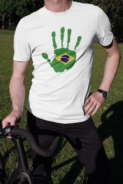 Camisa branca com estampa de uma mão verde e a bandeira do Brasil no centro, usada por um homem em um ambiente externo.