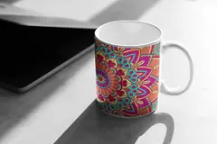 Caneca branca com design de mandala colorida nas cores rosa, azul e laranja, posicionada sobre uma superfície cinza.