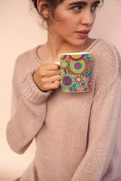 Caneca branca com estampa de mandala colorida, segurada por uma mulher com suéter rosa.