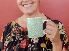 Caneca branca com design de mandala em fundo verde, segurada por uma mulher sorridente.