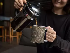 Uma mulher despejando café em uma caneca branca com design de mandala.