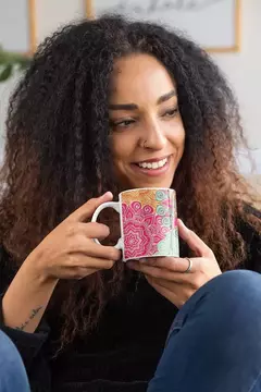 Caneca branca com estampa de mandala colorida, sendo segurada por uma mulher sorridente.