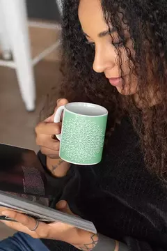 Mulher segurando uma caneca branca com desenho de mandala verde enquanto lê um livro.