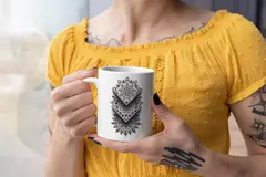 Caneca branca com design de mandala em preto, segurada por uma mulher com blusa amarela e tatuagens visíveis.