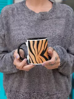 Caneca de porcelana com estampa de animal print em tons de laranja e preto, segurada por uma mulher com suéter cinza.