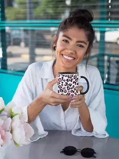 Mulher sorrindo enquanto segura uma caneca de porcelana com estampa animal print em uma mesa, ao lado de flores e óculos escuros.