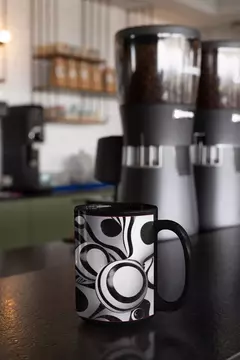 Caneca preta com design abstrato em bolas brancas e pretas, posicionada em uma mesa de café.