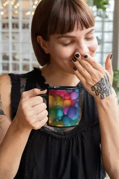 Caneca com design de bolas coloridas em fundo preto, sendo segurada por uma mulher com cabelo curto e tatuagens, sorrindo.