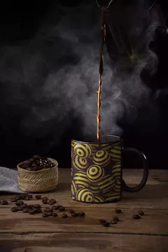 Caneca moderna com design abstrato em bolinhas amarelas e pretas, sendo servida com café quente, acompanhada de grãos de café e uma toalha de mesa.