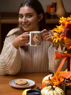 Mulher segurando uma caneca moderna com design abstrato colorido, sorrindo ao lado de uma mesa decorada com flores e abóboras.