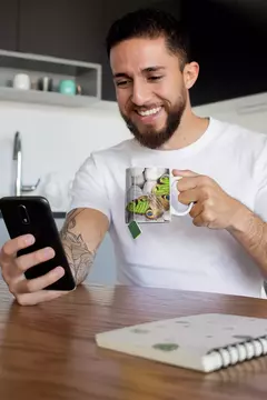 Homem sorridente segurando uma caneca com design de borboleta enquanto usa um celular.