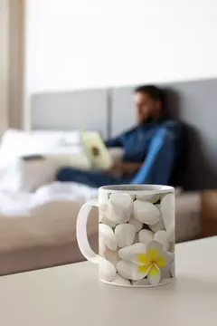 Caneca moderna com design de pedras brancas e uma flor amarela, em uma mesa ao lado de uma pessoa lendo.