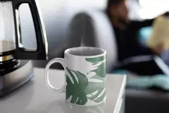 Caneca branca com estampa de folhagens verdes, fumegante, sobre uma mesa ao lado de uma cafeteira.