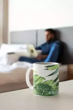 Caneca moderna com estampas de folhagens verdes, em primeiro plano, sobre uma mesa, enquanto um homem está desfocado ao fundo.