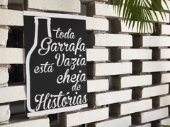 Quadro MDF com a frase "toda Garrafa Vazia está cheia de Histórias" em fundo preto, pendurado em uma parede de tijolos brancos.