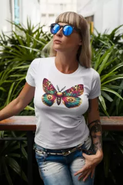 Modelo usando uma camisa branca BabyLook com estampa de borboleta colorida, em um ambiente ao ar livre com plantas ao fundo.