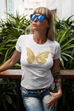 Modelo vestindo uma camisa branca BabyLook com estampa de borboleta dourada, em meio a plantas verdes.