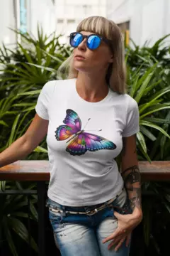 Modelo usando uma camisa branca BabyLook com estampa de borboleta colorida, em um ambiente com plantas ao fundo.